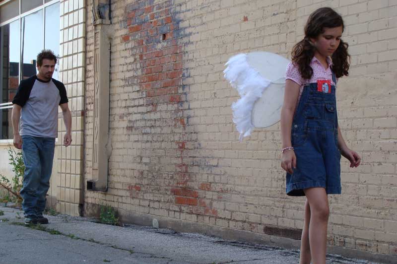 Man follows young girl wearing angel wings down a dingy alley